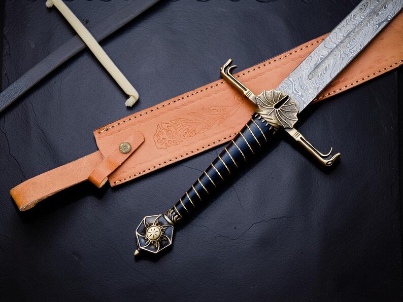 Op de afbeelding: Een zwaard met een patroon van Damascusstaal, een zwart handvat omwikkeld met gouddraad en een messing stootplaat. De pommel heeft een zonnestraalontwerp. Het wordt vergezeld door een beige lederen schede met in reli&euml;f aangebrachte bloemdetails.