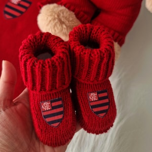 Pode incluir: Um conjunto de bebé de malha vermelha com chapéu, camisola, macacão e botinhas. O conjunto inclui um ursinho de peluche com macacão. Cada artigo tem um brasão com um design vermelho e azul marinho.