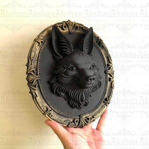 Puede incluir: Decoración de pared con cabeza de zorro negro en un marco ovalado con adornos dorados. El zorro está esculpido en detalle, con pelaje visible y orejas puntiagudas. El marco tiene una estética vintage.