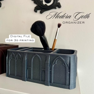 Puede incluir: Un organizador gris oscuro, impreso en 3D, con un diseño de arco gótico, que contiene brochas de maquillaje. El texto "Modern Goth ORGANIZER" y "DIGITAL FILE FOR 3D PRINTING" son visibles. El organizador está sobre una superficie blanca, con detalles decorativos negros en el fondo.