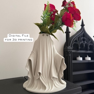 May include: A cream-colored ghost-shaped vase holding red roses and green foliage. The vase has a draped design and three eye-shaped holes. Text on the image reads "DIGITAL FILE FOR 3D PRINTING". The vase is on a black surface.