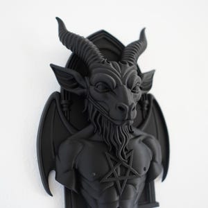 May include: A black wall sculpture of Baphomet, a goat-headed figure with horns, wings, and a pentagram on its chest. The sculpture is mounted on a wall and has a detailed, three-dimensional design. The sculpture is a gothic or occult-themed decorative item.