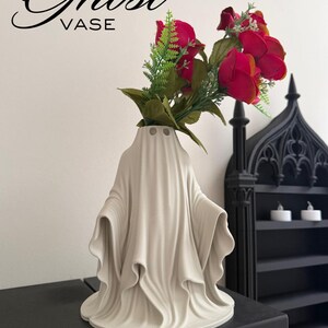 May include: A white ceramic ghost-shaped vase with the word "Ghost VASE" in black script. The vase holds red roses and green foliage. The vase has a draped design, and is on a black surface.