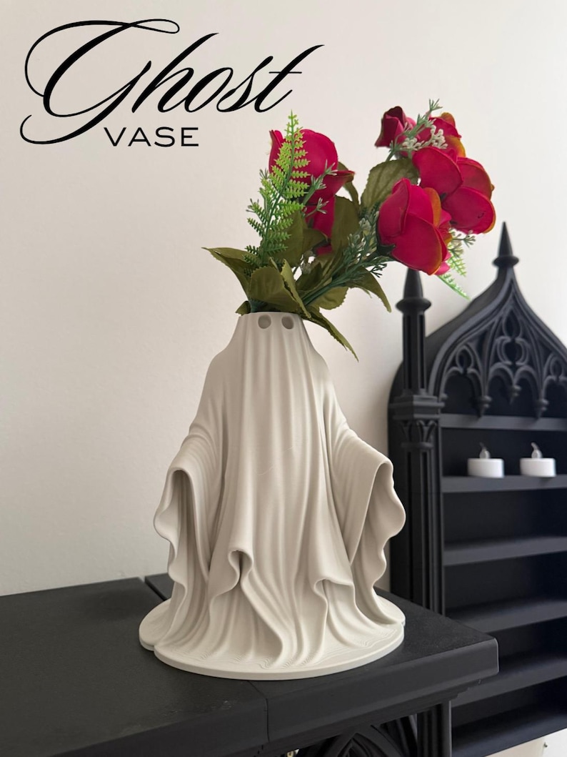 May include: A white ceramic ghost-shaped vase with the word "Ghost VASE" in black script. The vase holds red roses and green foliage. The vase has a draped design, and is on a black surface.
