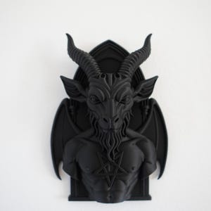 May include: A black wall sculpture depicting Baphomet, a goat-headed deity, with large horns, wings, and a pentagram on its chest. The sculpture is mounted on a black, arched background, and is set against a white wall.
