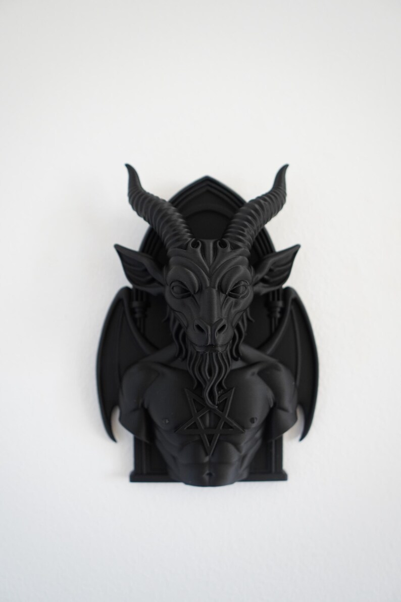 May include: A black wall sculpture depicting Baphomet, a goat-headed deity, with large horns, wings, and a pentagram on its chest. The sculpture is mounted on a black, arched background, and is set against a white wall.