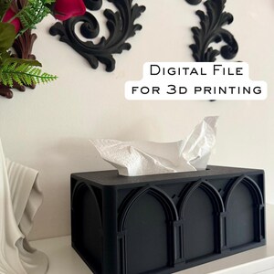 May include: A black tissue box with an arched design, holding white tissues. The box is rectangular and sits on a white shelf. The text "DIGITAL FILE FOR 3D PRINTING" is visible. Decorative black wall accents and red flowers are in the background.