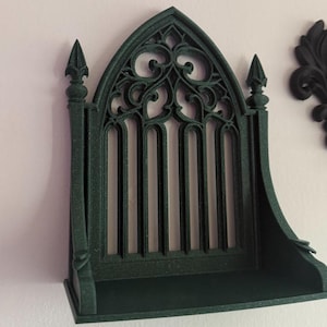 Peut inclure: &Eacute;tag&egrave;re murale de style gothique vert fonc&eacute; avec un sommet en arc et des barres verticales. L'&eacute;tag&egrave;re a une petite corniche et est mont&eacute;e sur un mur blanc. Des &eacute;l&eacute;ments d&eacute;coratifs sont visibles sur le dessus et les c&ocirc;t&eacute;s.