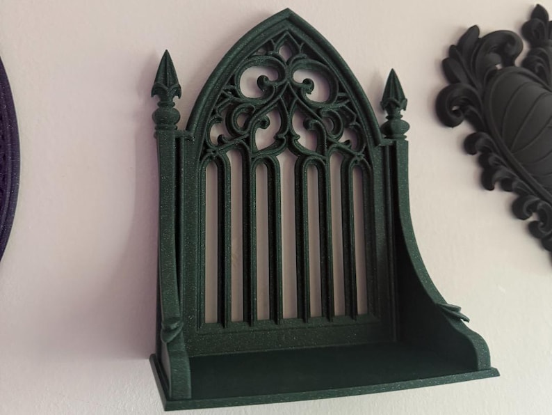 Peut inclure: &Eacute;tag&egrave;re murale de style gothique vert fonc&eacute; avec un sommet en arc et des barres verticales. L'&eacute;tag&egrave;re a une petite corniche et est mont&eacute;e sur un mur blanc. Des &eacute;l&eacute;ments d&eacute;coratifs sont visibles sur le dessus et les c&ocirc;t&eacute;s.
