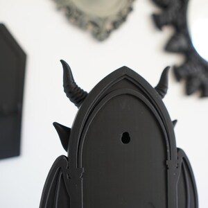 May include: Black gothic-style wall decor featuring a horned devil design. The decor has a pointed arch shape with bat wings and a hole for hanging. Other black gothic mirrors and a coffin-shaped item are in the background.