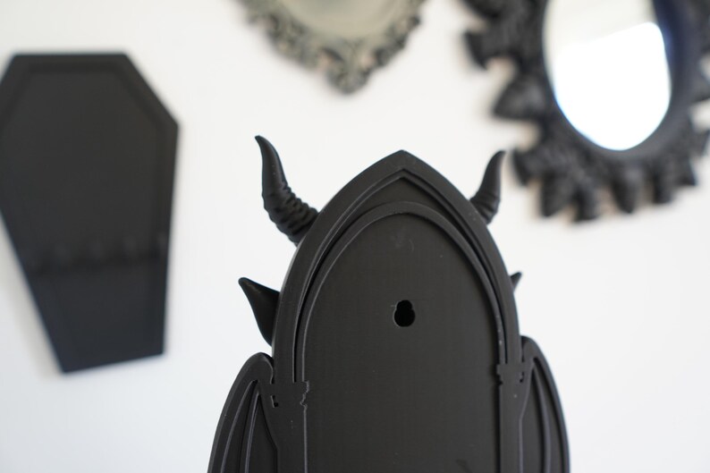 May include: Black gothic-style wall decor featuring a horned devil design. The decor has a pointed arch shape with bat wings and a hole for hanging. Other black gothic mirrors and a coffin-shaped item are in the background.