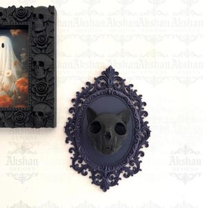 May include: A black cat skull wall decor piece mounted on a dark blue oval frame with ornate detailing. The skull is centered within the frame, creating a gothic or Halloween theme. The background is white.