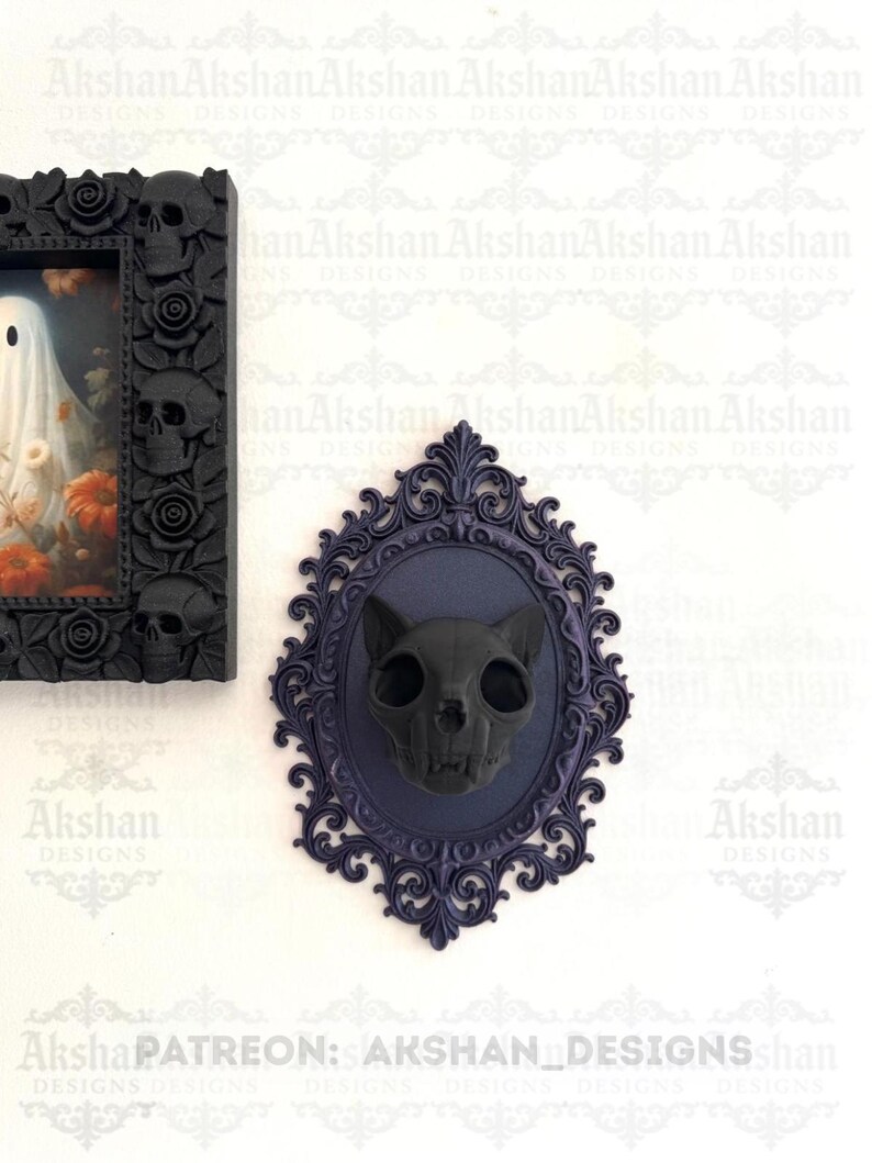 May include: A black cat skull wall decor piece mounted on a dark blue oval frame with ornate detailing. The skull is centered within the frame, creating a gothic or Halloween theme. The background is white.
