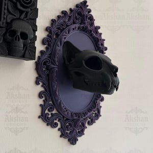 May include: A black cat skull mounted on a purple oval plaque with ornate, dark purple trim. The plaque is mounted on a white wall. The image also shows a black skull and rose design on a black frame. Text on the image reads "PATREON: AKSHAN_DESIGNS."