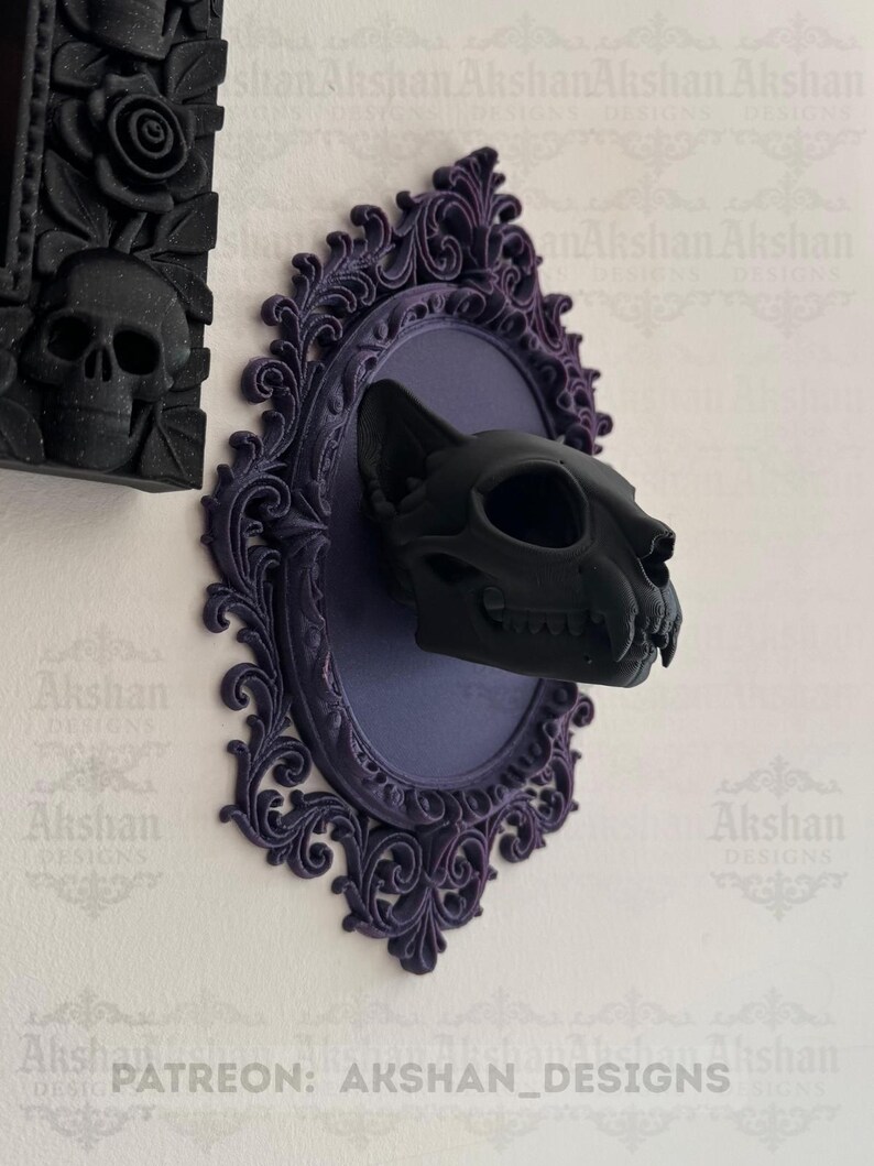 May include: A black cat skull mounted on a purple oval plaque with ornate, dark purple trim. The plaque is mounted on a white wall. The image also shows a black skull and rose design on a black frame. Text on the image reads "PATREON: AKSHAN_DESIGNS."