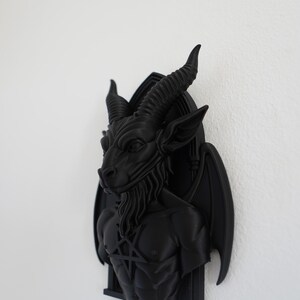 May include: A black wall sculpture of Baphomet, a goat-headed deity, with large horns, a beard, and a pentagram on its chest. The sculpture has bat-like wings and is mounted on a black frame. The artwork is a gothic or occult-themed decorative item.