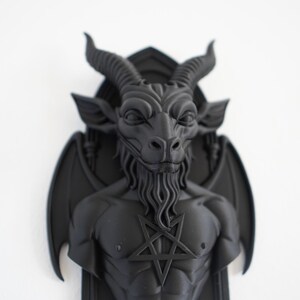 May include: A black wall sculpture depicting a horned, bearded figure with a pentagram on its chest. The sculpture has bat-like wings and is mounted on a rectangular base. The figure's face has a stern expression.