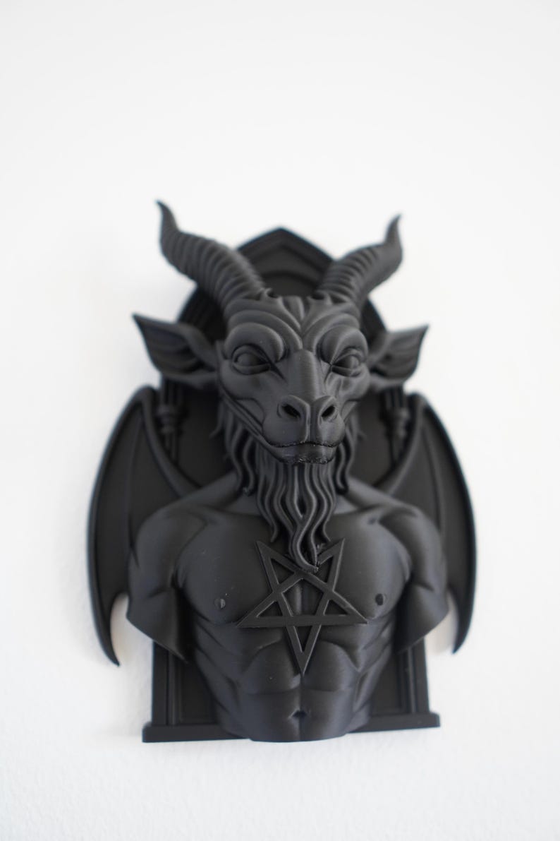 May include: A black wall sculpture depicting a horned, bearded figure with a pentagram on its chest. The sculpture has bat-like wings and is mounted on a rectangular base. The figure's face has a stern expression.