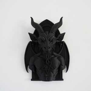 May include: A black wall sculpture depicting Baphomet, a goat-headed deity, with large horns, wings, and a pentagram on its chest. The sculpture is mounted on a white wall, showcasing the detailed features of the dark artwork.