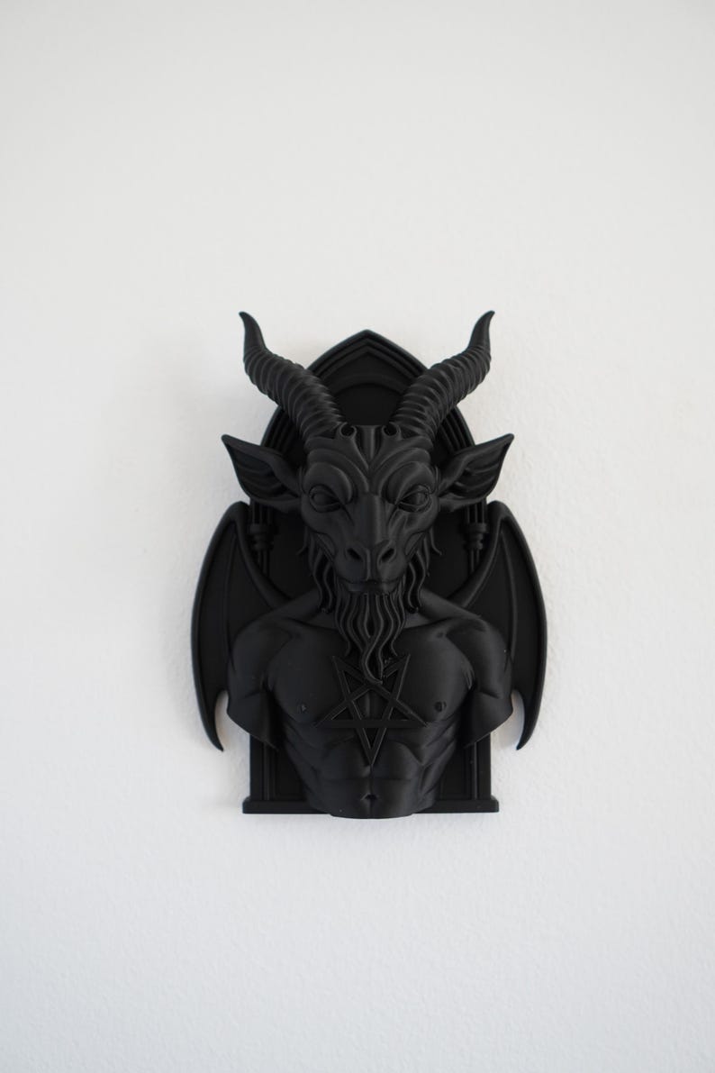 May include: A black wall sculpture depicting Baphomet, a goat-headed deity, with large horns, wings, and a pentagram on its chest. The sculpture is mounted on a white wall, showcasing the detailed features of the dark artwork.