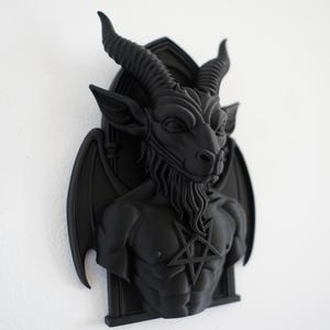 May include: A black wall sculpture of Baphomet, a goat-headed figure with horns, wings, and a pentagram on its chest. The sculpture is mounted on a white wall, showcasing the detailed features of the dark artwork.