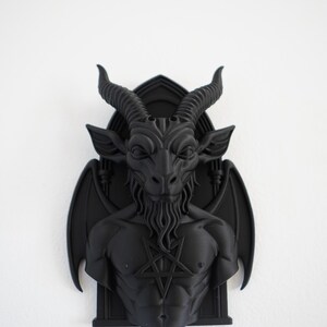 May include: A black wall sculpture depicting Baphomet, a goat-headed deity, with a pentagram on its chest. The sculpture features detailed horns, a beard, and wings, mounted on a gothic-style frame. The artwork is a dark, gothic-themed decorative piece.
