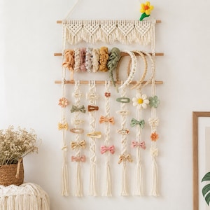 May include: A cream-colored macrame wall hanging organizer featuring three horizontal wooden dowels. The organizer holds various fabric scrunchies, headbands, and numerous hair clips in shades of pink, green, yellow, and white. A small yellow plush flower is attached to the top right corner.