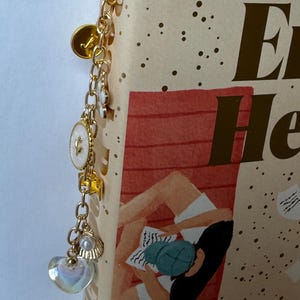 May include: Gold-toned bookmark with a chain of charms, including a heart, pearl, and a white oval with a leaf design. The bookmark is attached to a book with the title "Every Heart a Doorway".