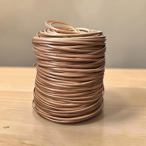 May include: A cylindrical spool of light brown leather cord wound in tight, uniform layers. The cord has a smooth, rounded texture and a consistent tan color throughout the entire coil, creating a neat, stacked appearance of thin, flexible material.