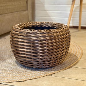 May include: A circular wicker basket in shades of brown. The basket has a cylindrical shape and is placed on a natural fibre rug. The basket is empty and suitable for storage or decorative purposes.
