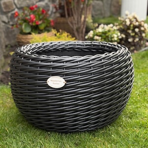 Può includere: Un vaso rotondo nero intrecciato, posto sull'erba verde. Il vaso ha un'ampia apertura e un'etichetta con la scritta "Maysternya Kashpo". Il vaso è realizzato in materiale scuro ed è circondato da verde e fiori.