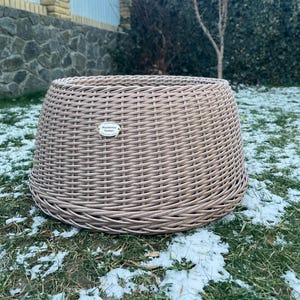 Może przedstawiać: Tkany, jasnobrązowy kosz zewnętrzny o szerokim, zaokrąglonym kształcie. Kosz wykonany jest z ciasno tkanej tkaniny i ma małą, prostokątną metkę przymocowaną z przodu. Kosz stoi na trawie z płatami śniegu.