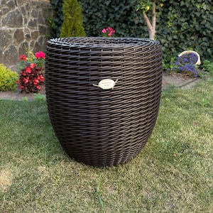 May include: A dark brown, woven wicker planter with a rounded, tapered shape. The planter sits on a grassy lawn, with a backdrop of greenery and flowers. A small, oval tag is attached to the planter.