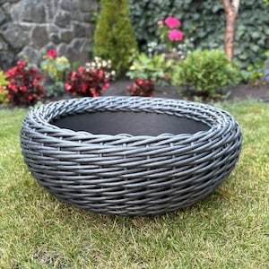 Può includere: Una fioriera grigia, in stile intrecciato, di forma rotonda. La fioriera è posizionata su un letto di erba verde e ha un interno scuro. L'esterno è realizzato in un materiale grigio simile al vimini. Lo sfondo presenta un giardino con fiori e vegetazione.