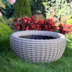 Könnte beinhalten: Ein grauer Pflanzkübel im Webstil steht auf grünem Gras. Der runde Pflanzkübel hat einen dunklen Innenraum und ist von einem Garten mit roten Blumen und grünem Laub umgeben. Der Pflanzkübel besteht aus einem haltbaren, wetterbeständigen Material.