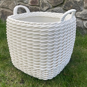 May include: A white, woven basket with two curved handles. The basket is made of a thick, textured material and sits on green grass. The background features a stone wall.