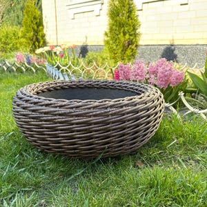 May include: A round, woven wicker planter with a dark interior sits on green grass. The planter is brown and has a textured surface. The background includes a house, flowers, and trees.