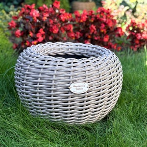May include: A round, gray woven planter basket sits on green grass. The basket has a woven design and a small, oval tag with text. Red flowers and green foliage are in the background. The planter is designed for outdoor use.