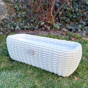 Pode incluir: Uma floreira retangular branca de vime com um interior liso e de cor clara. A floreira está colocada sobre relva verde, com uma parede de pedra e folhagem no fundo. Uma pequena etiqueta decorativa está presa na frente.