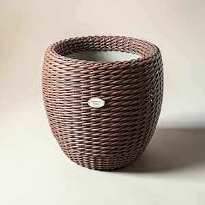 May include: A brown woven wicker planter with a rounded shape. The planter has a wide opening and a label on the front. The woven texture is visible throughout the planter's exterior. The interior appears to be a lighter color.