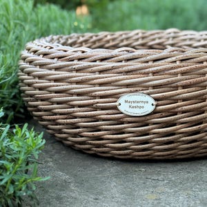 Pode incluir: Uma cesta redonda tecida em tons naturais. A cesta é feita de tiras entrelaçadas de cor marrom claro, criando uma superfície texturizada. Uma pequena etiqueta oval com texto está anexada. A cesta está sobre uma superfície cinza com folhagem verde ao fundo.