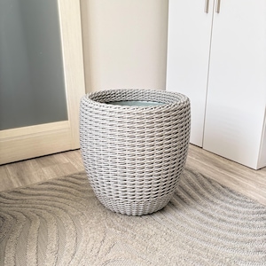May include: A light gray woven basket-style planter. The planter has a rounded shape and is sitting on a light gray rug with a wave pattern. The background includes a mirror and a white cabinet.