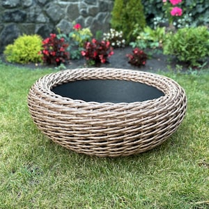 May include: A round, woven planter with a dark interior sits on a grassy lawn. The planter is made of light brown wicker and has a low profile. The background features a stone wall, flowers, and greenery.