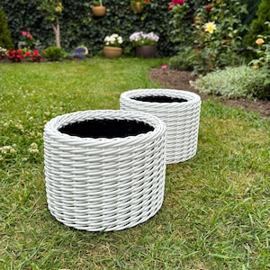 Pode incluir: Duas floreiras redondas brancas tecidas com interiores pretos estão sobre um gramado verde. As floreiras são cilíndricas e têm uma aparência texturizada, semelhante a uma cesta. O fundo apresenta um jardim exuberante.