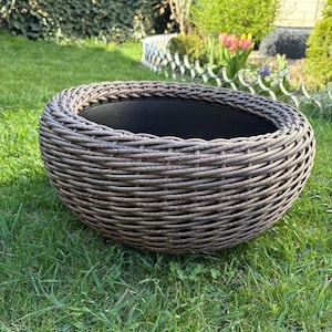 Op de afbeelding: Een ronde, lage plantenbak met een gevlochten, donkerbruine buitenkant van synthetisch vlechtwerk en een gladde, effen zwarte binnenbak. Het ontwerp heeft een dikke, afgeronde rand en een brede, komvormige structuur voor planten.