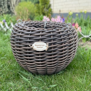 Puede incluir: Una cesta redonda de mimbre marrón con una etiqueta que dice "Maysternya Kashpo". La cesta está hecha de tiras entrelazadas, creando una superficie texturizada. Se encuentra sobre hierba verde, con un fondo borroso de plantas y flores.