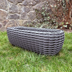 May include: A gray, rectangular woven planter sits on green grass. The planter has a rounded shape and is made of a woven material. The background features a stone wall and some greenery.