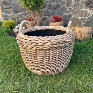 May include: A large, round, woven wicker basket with two curved handles. The basket is light brown and sits on green grass. The interior of the basket is black. Other wicker baskets and plants are in the background.