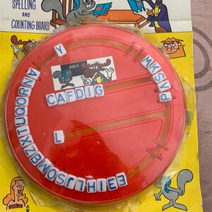 May include: A red plastic Bullwinkle's Spelling and Counting Board game with a rotating dial featuring letters of the alphabet. The game includes a small plastic moose figure and a small plastic squirrel figure. The game is priced at 69 cents.