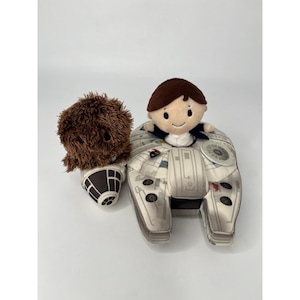May include: Plush toys featuring characters from a science fiction franchise. One plush is a brown, furry creature. Another is a character with brown hair and a blue jacket, seated in a Millennium Falcon-shaped plush.